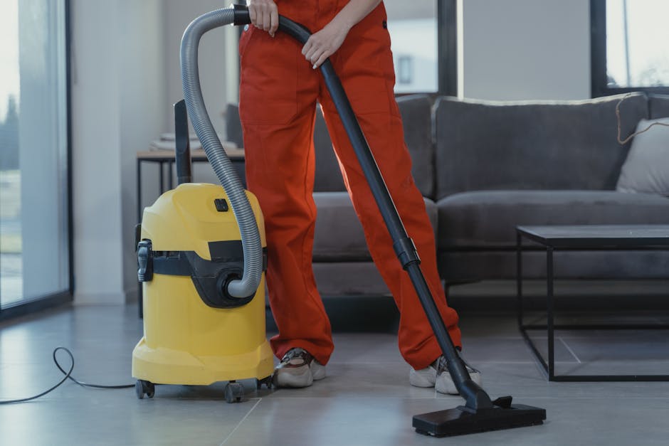 A woman vacuuming a patterned area rug on a wooden floor in a living room, with a brown leather sofa partially visible on the right side. The rug features intricate floral designs in muted tones, and 