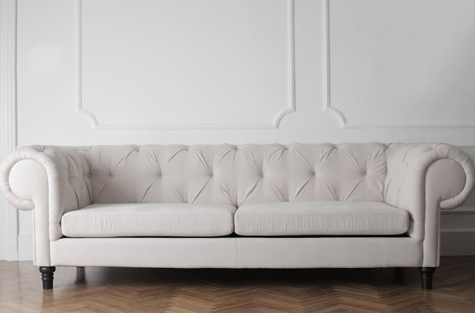 A pristine white upholstered sofa with tufted cushioning and rounded armrests, situated against a decorated white wall with panel molding. The sofa is positioned on a wooden floor, and the room is wel