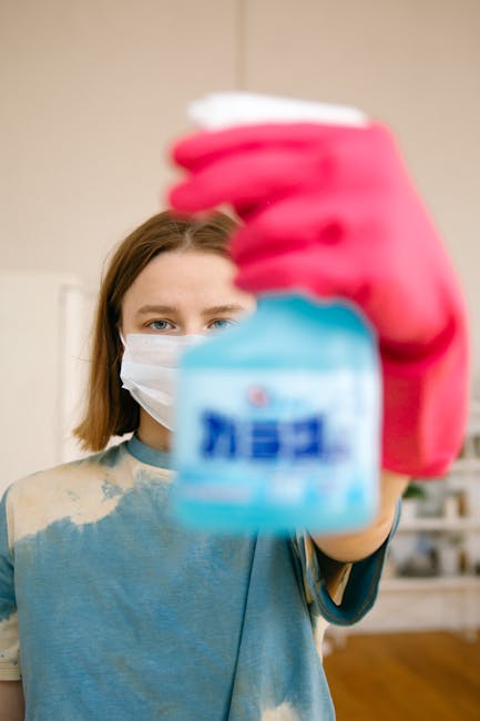 A person dressed in full protective gear, including a white hazmat suit, face mask, and blue gloves and shoe covers, is performing deep cleaning on a beige carpet in a residential living room. The ind